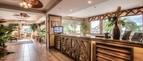 A hotel reception area with a wooden reception desk, ceiling fans, and large windows offering a view of palm trees.