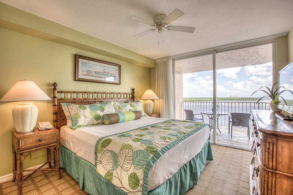 A bedroom with a bed, bedside lamps, a ceiling fan, a large window, and a view of the outdoors.