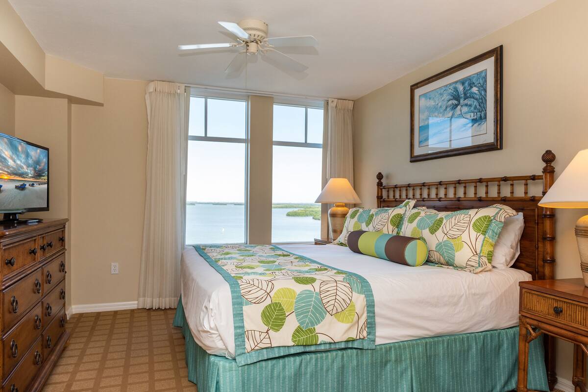 A bedroom with a bed, a television, a ceiling fan, a window with curtains, and a painting on the wall.