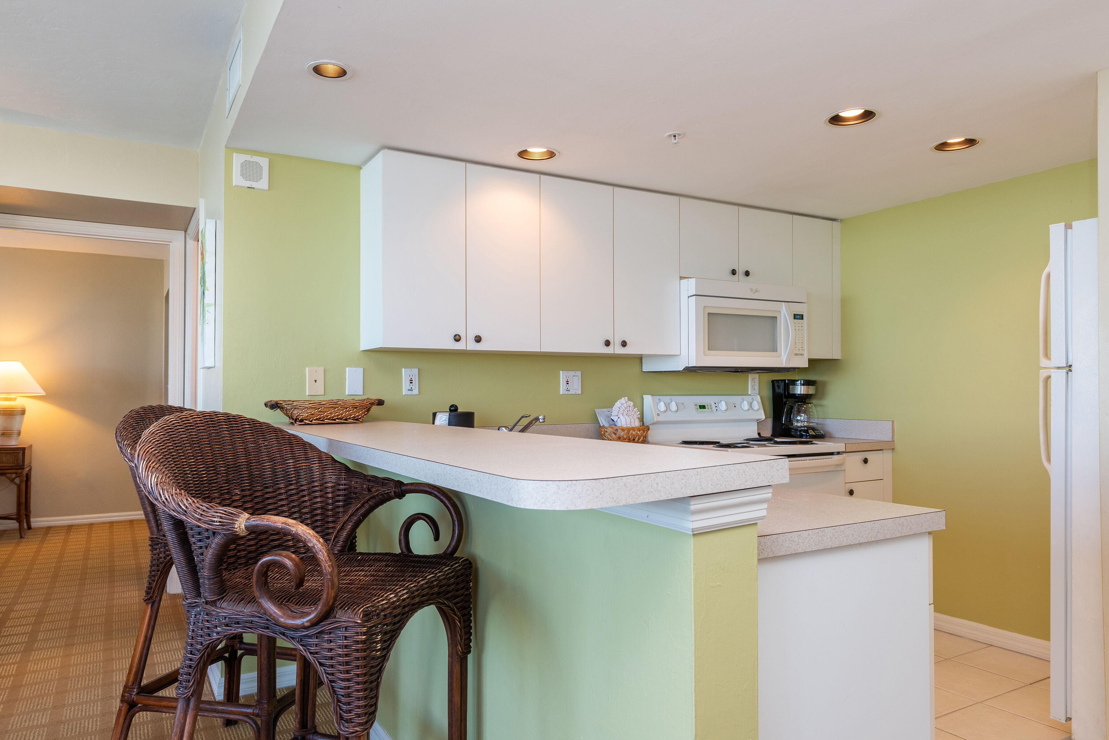 A kitchen with a white microwave, stove, and white cabinets. There is a wicker chair and a small island with a sink.