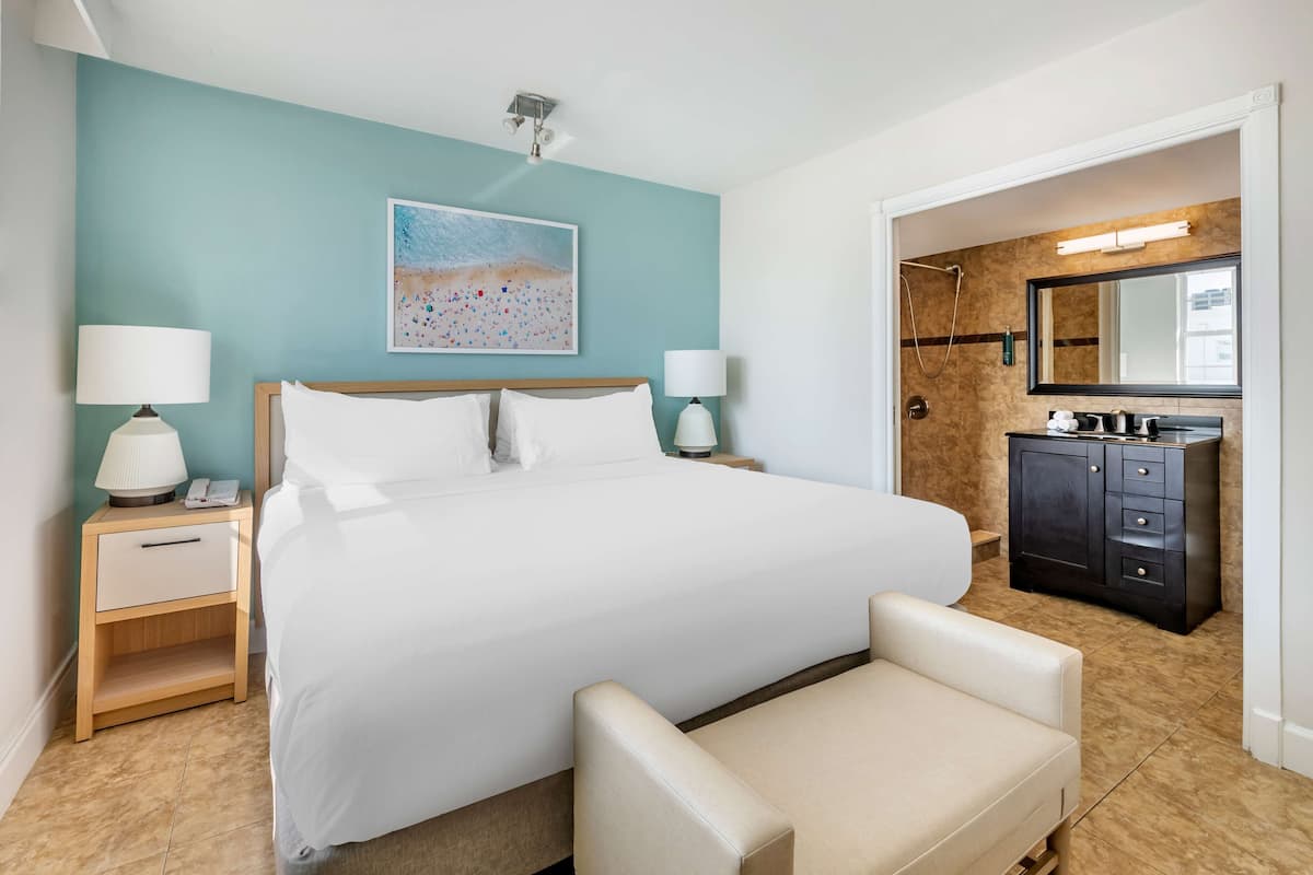 Une chambre d’hôtel avec un grand lit, des lampes de chevet, une table de chevet, un banc, une salle de bain avec un miroir, un lavabo et un meuble de rangement.