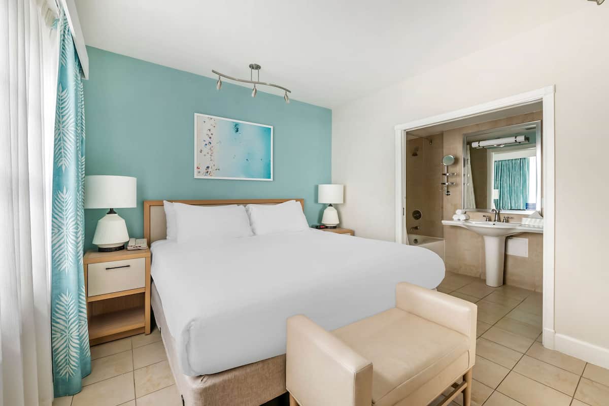 Une chambre à coucher comprenant un lit, des tables de chevet, une commode, une chaise, une salle de bain avec un lavabo et un miroir, et un mur orné d’un tableau.