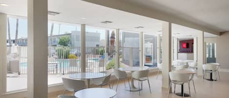 Un espacio interior amplio con varias mesas y sillas, y ventanas grandes que ofrecen vistas a una piscina exterior y edificios.