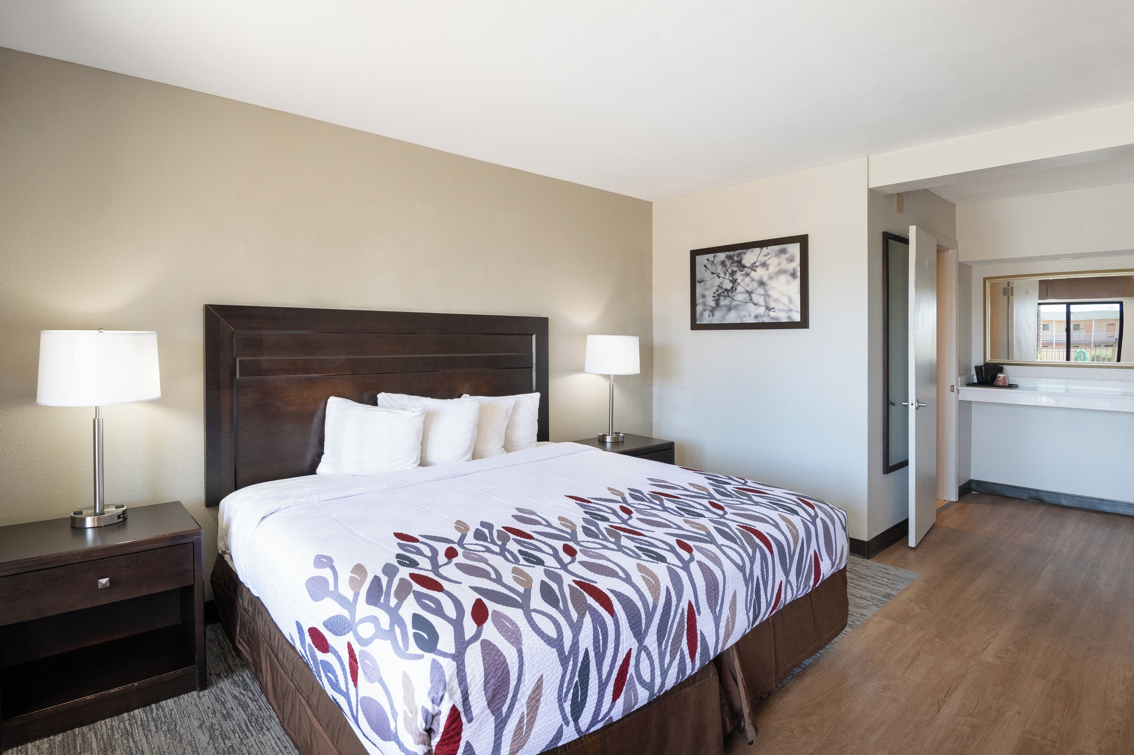 Habitación de hotel con una cama grande, dos lámparas de noche, una mesita de noche, una pequeña mesa, un cuadro en la pared y vistas de la habitación contigua.