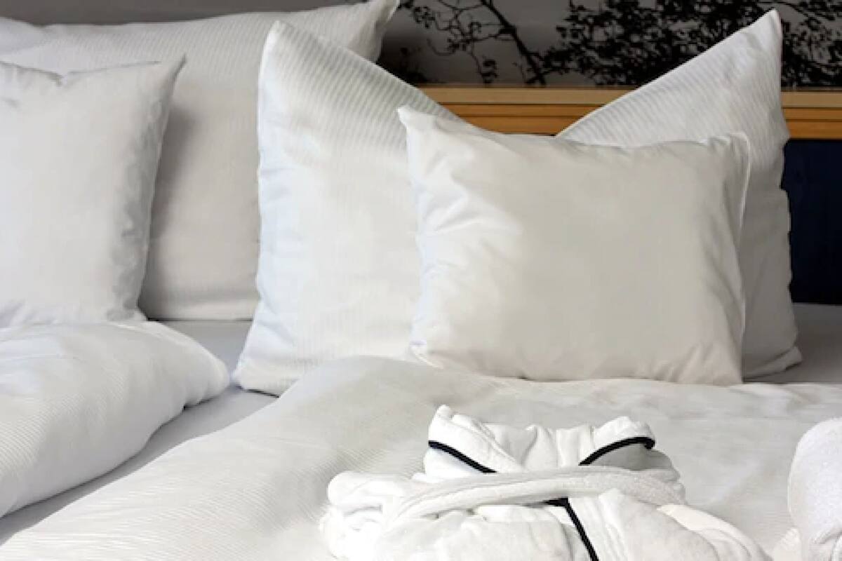 Una cama bien tendida con ropa y almohadas blancas, un cabecero de madera y una bata blanca doblada sobre la cama.
