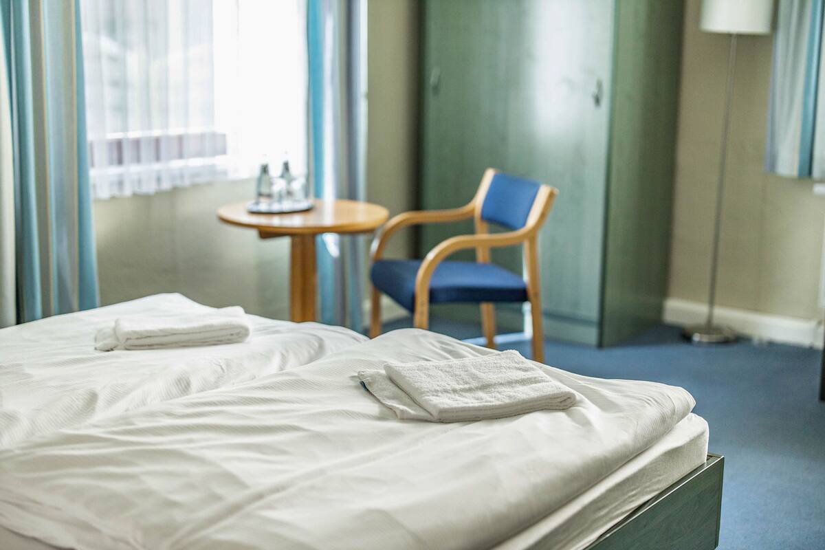 Una habitación de hotel con una cama bien hecha, una silla, una mesita con una botella y un vaso, y una lámpara.