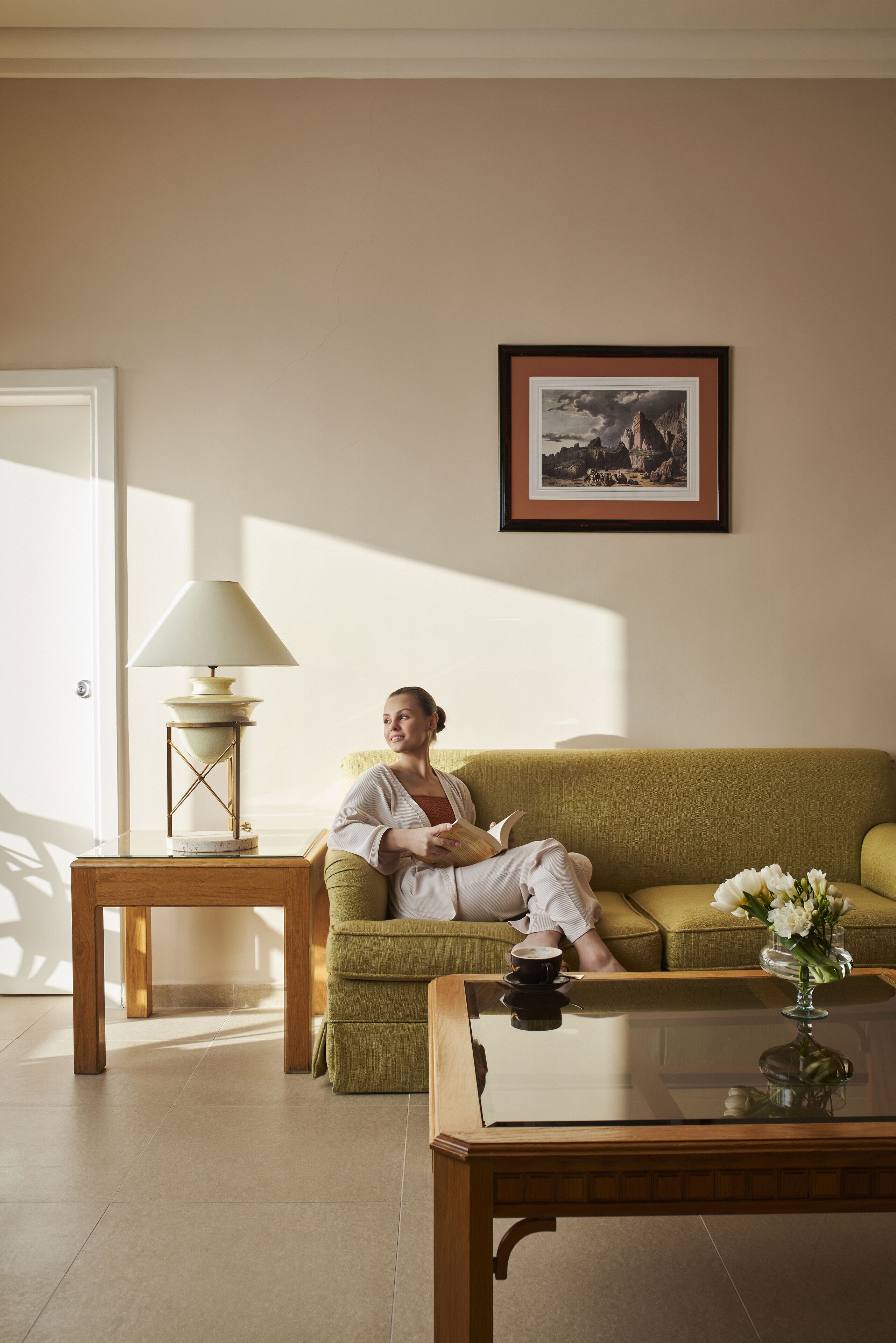 A room with a green sofa, a small wooden table with a lamp, a framed picture on the wall, and a vase with flowers on a coffee table.