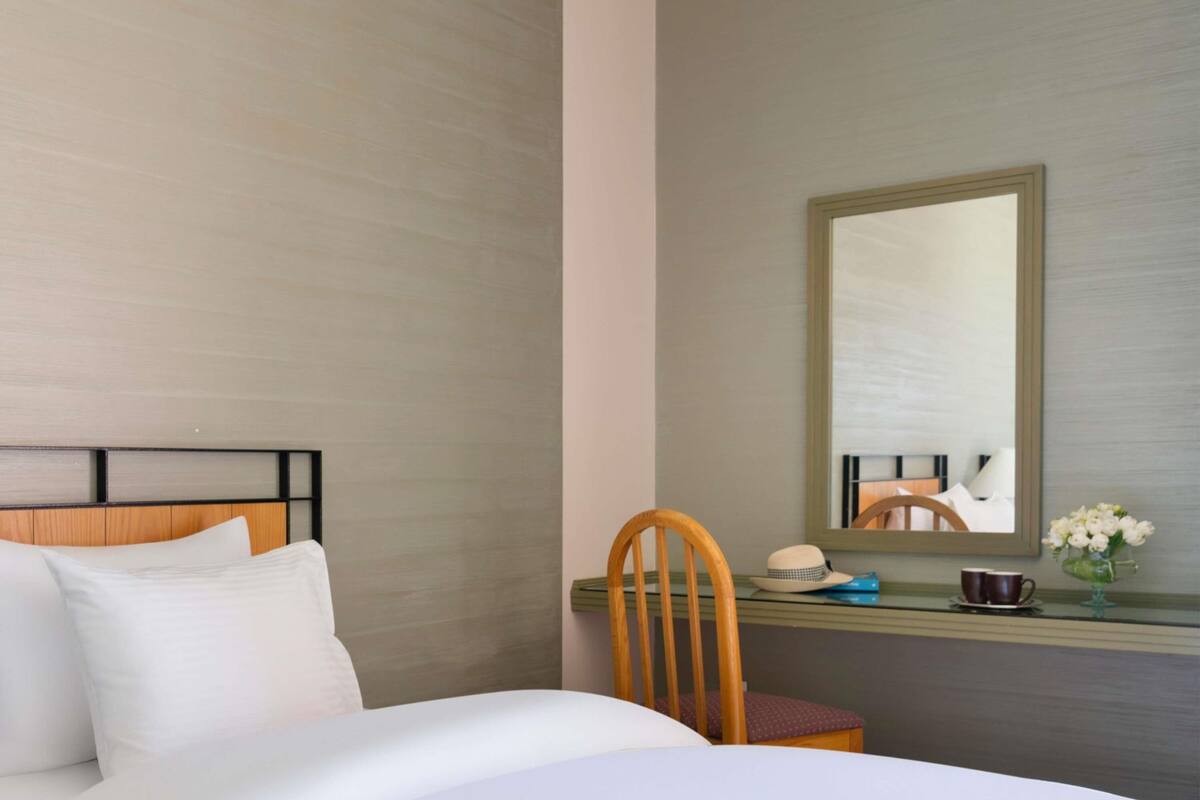 A hotel room with a bed, a wooden chair, a desk with a mirror, and a vase with flowers.
