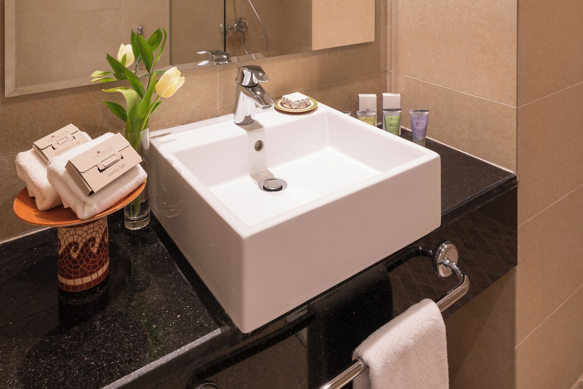 A modern bathroom with a large mirror, a white sink, and a shower. There are toiletries on the counter and a towel rack.