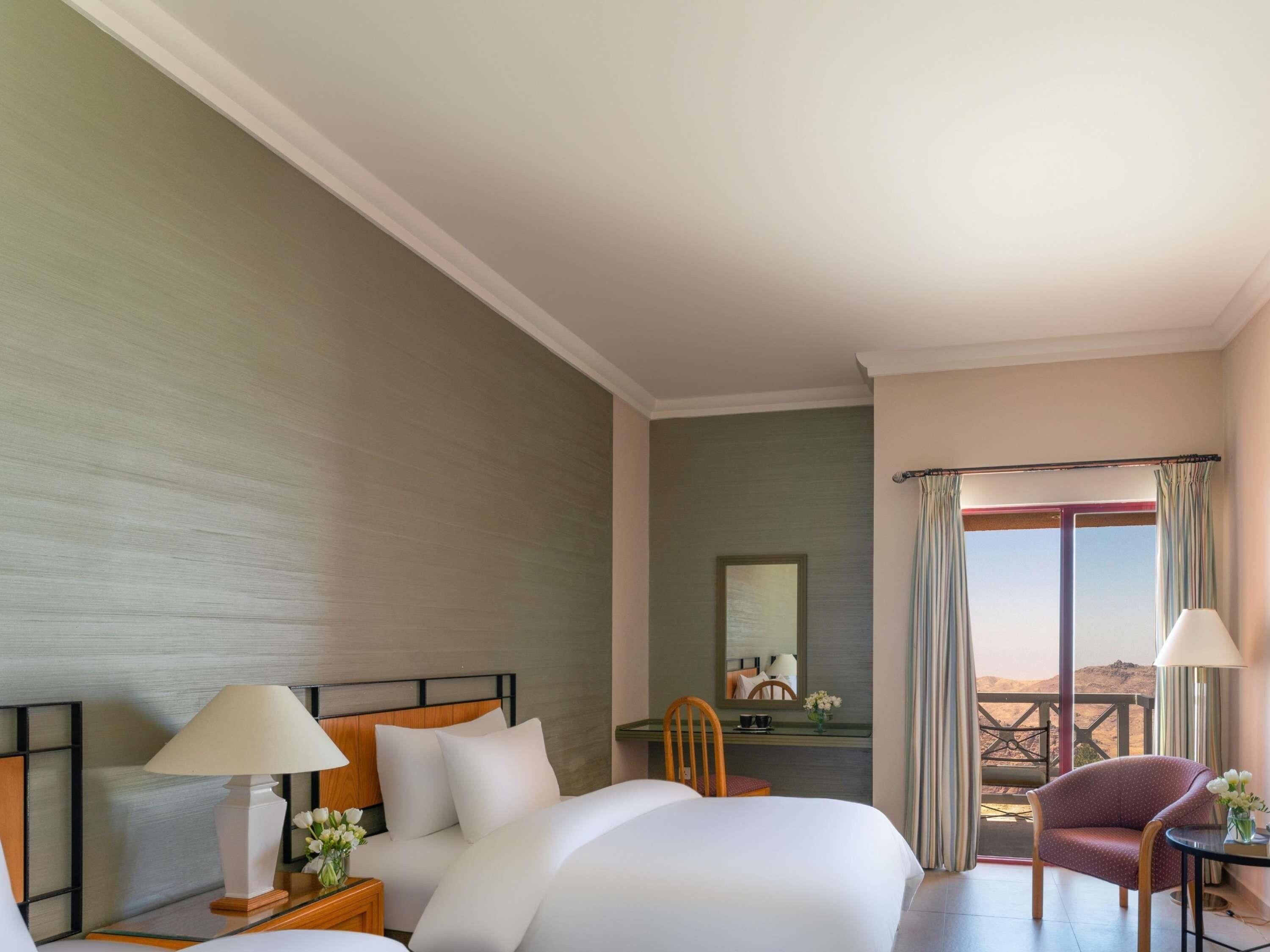 A hotel room with a large bed, a desk with a chair, a small table with a chair, and a balcony with curtains.