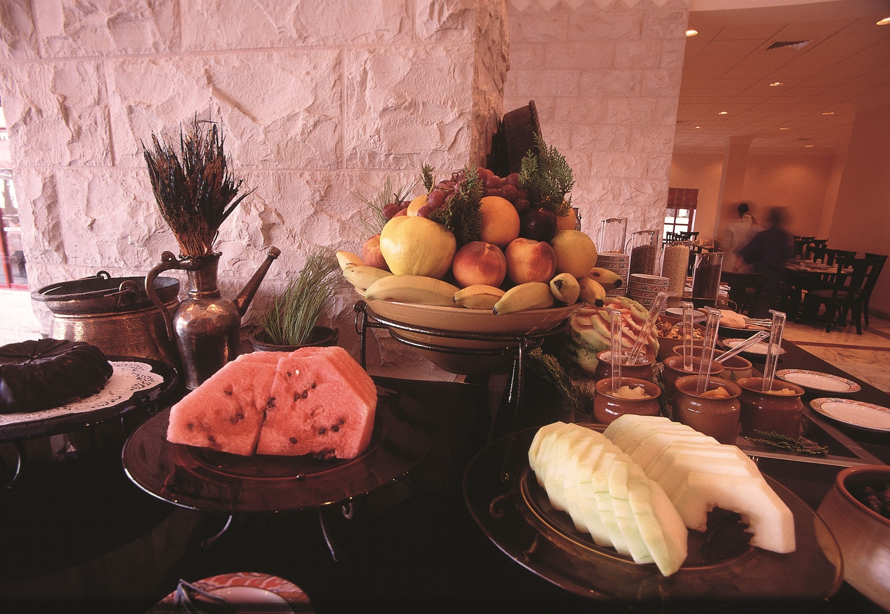 A buffet with a variety of fruits and desserts, including watermelon, bananas, and a cake, set against a stone wall background.