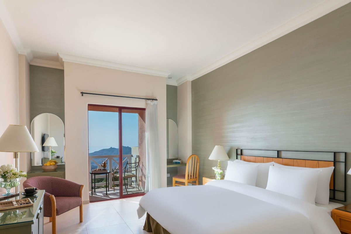 A hotel room with a large bed, a desk with a lamp, a chair, and a balcony with a view of the mountains.