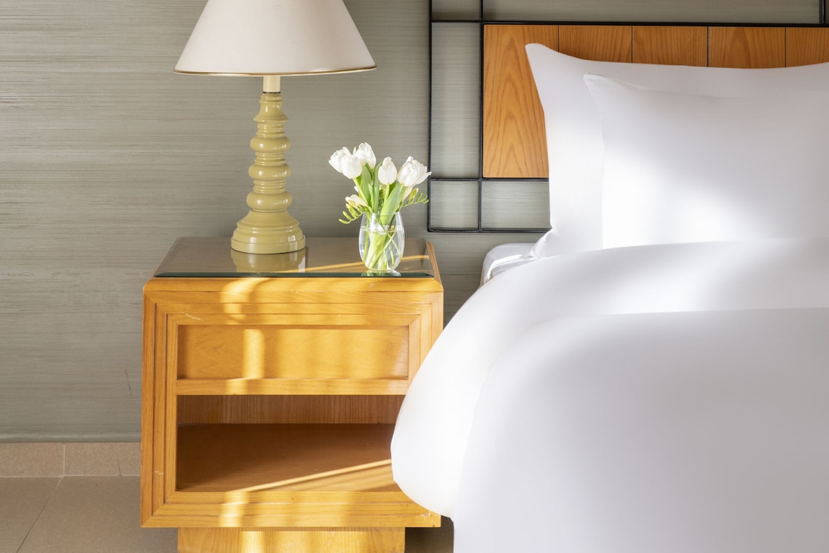 A neatly made bed with white linens, a wooden bedside table with a lamp, and a vase with flowers.