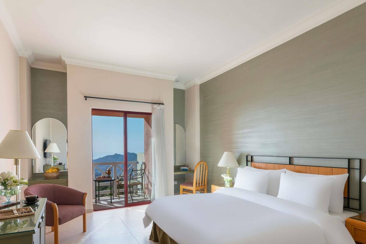 A hotel room with a large bed, a desk with a lamp, a chair, and a balcony with a view of the mountains.