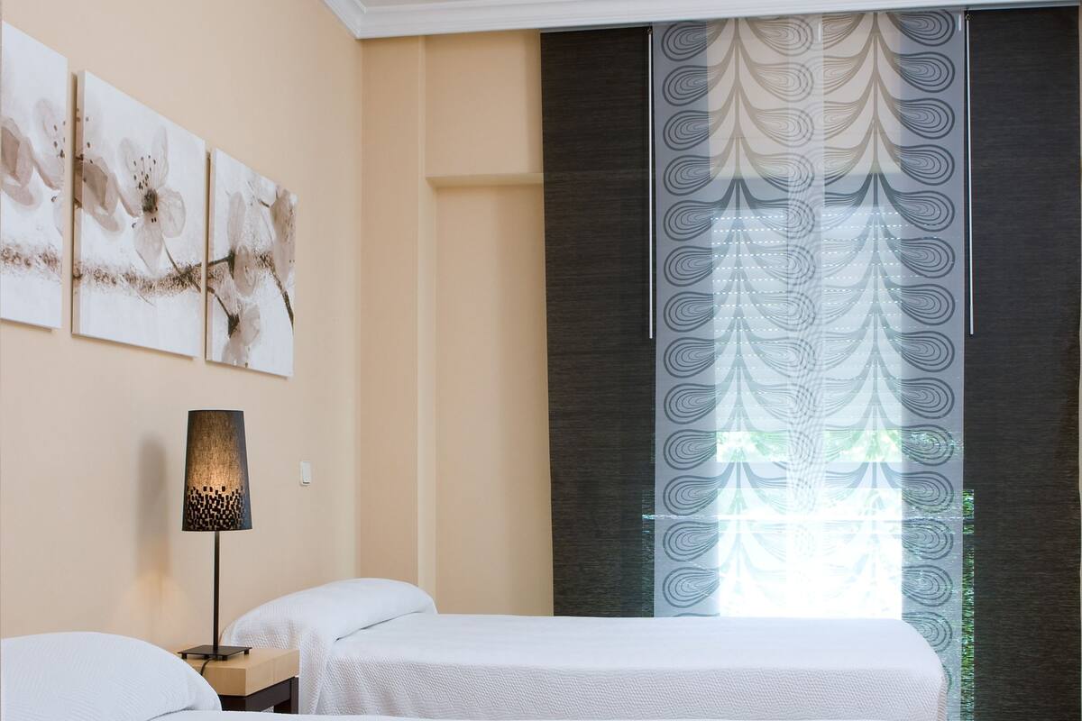 A hotel room with two beds, a lamp, and a window with patterned curtains.