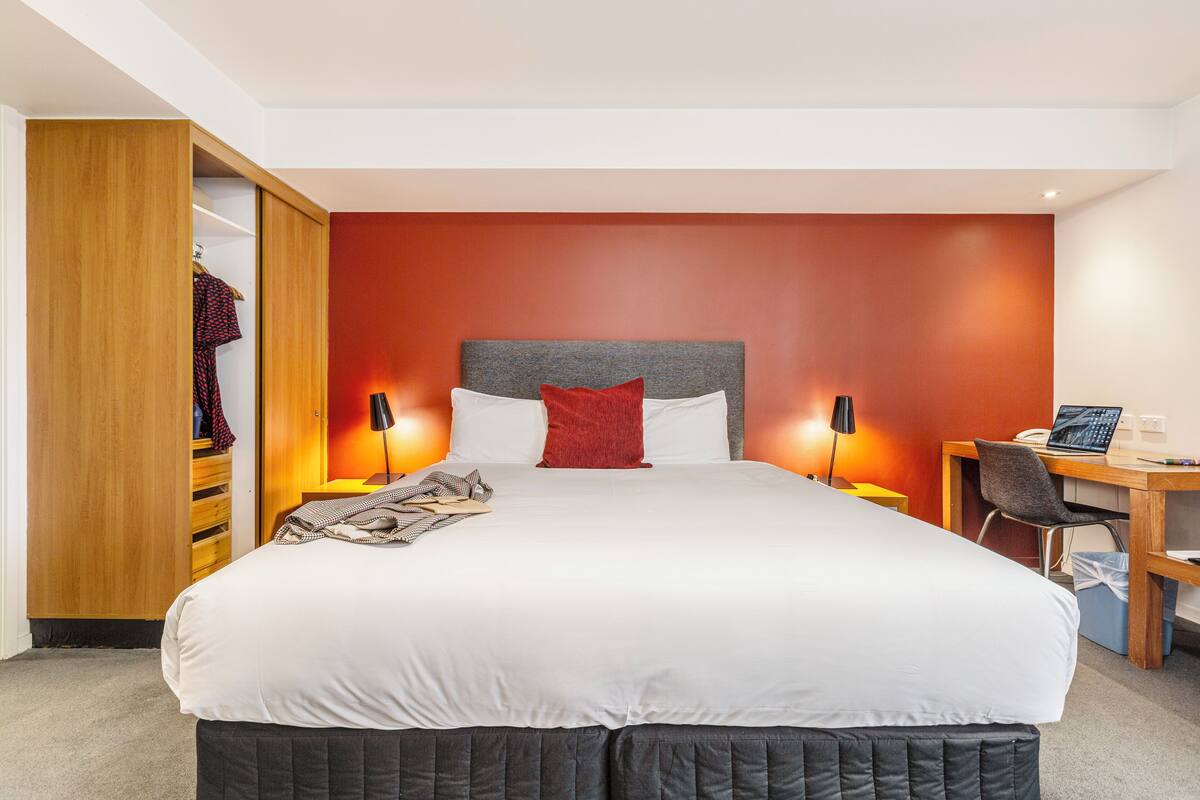 A hotel room with a large bed, a desk with a chair, a wardrobe, and a red accent wall.