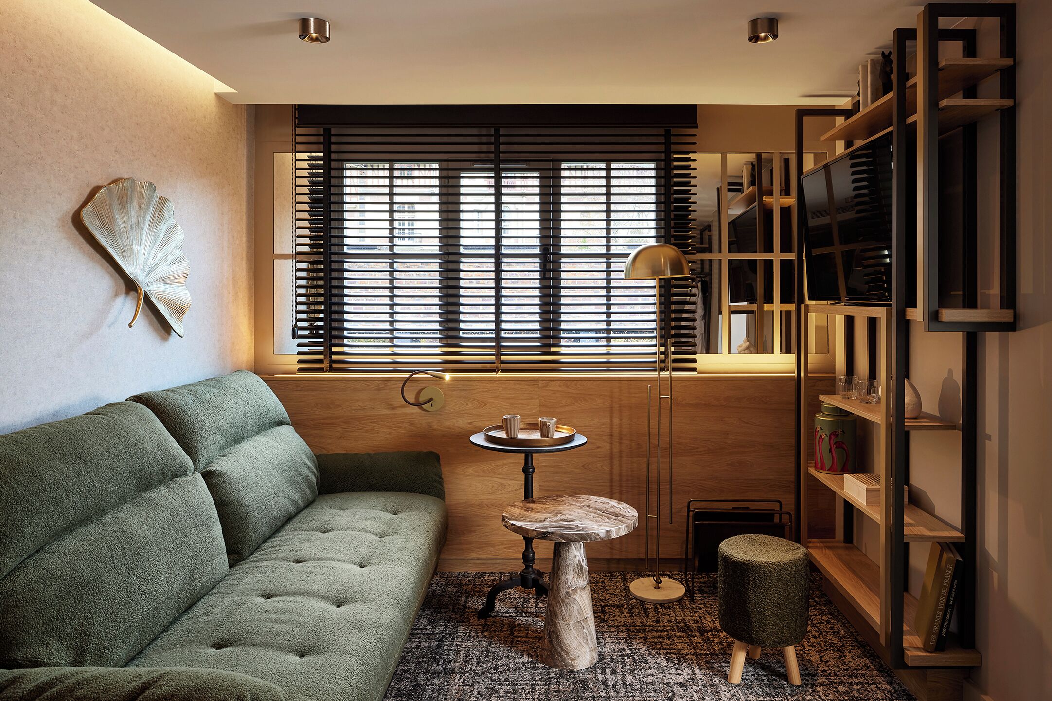 A cozy living room with a green sofa, a round wooden table, a bookshelf, and a large window with blinds.