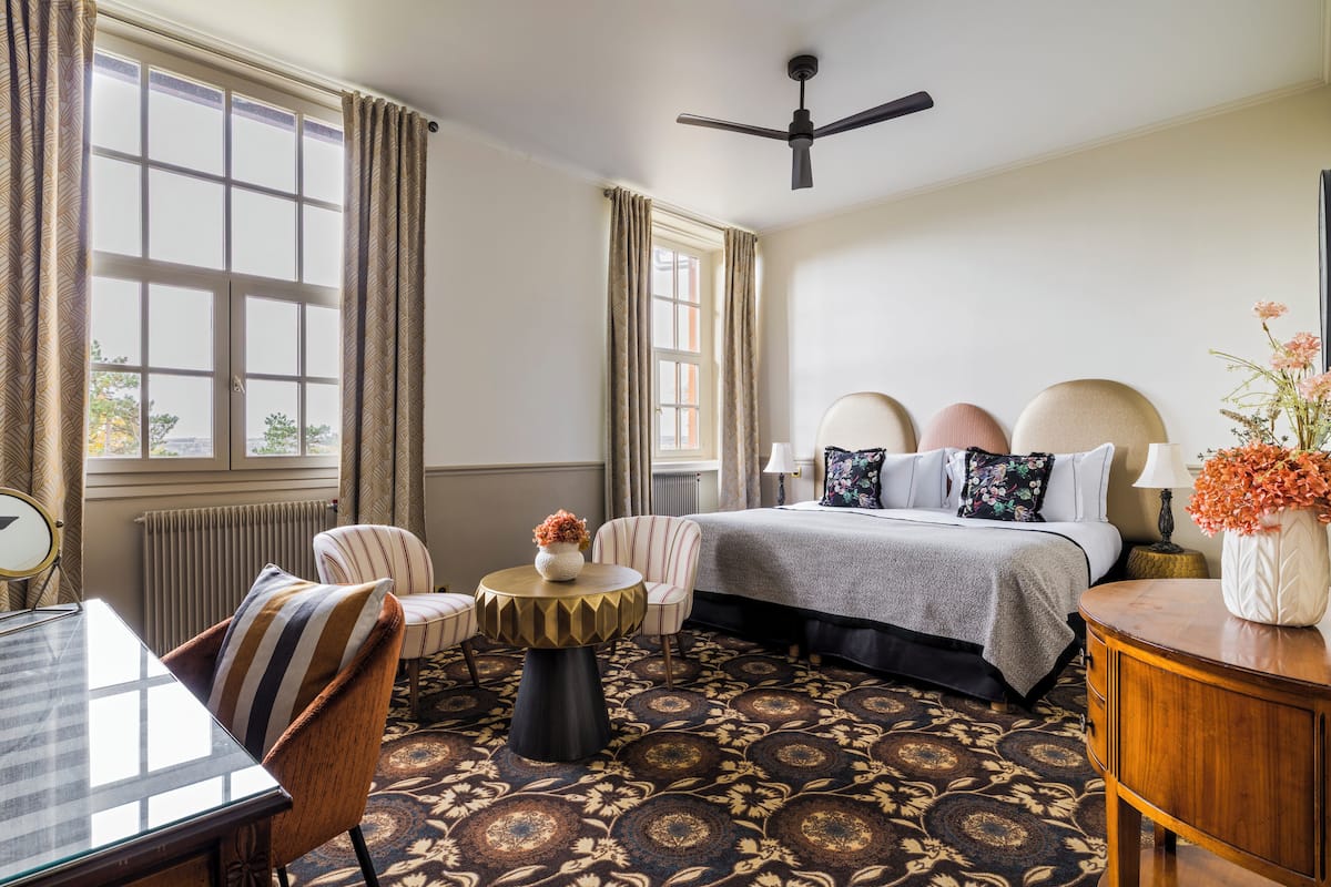 A hotel room with a bed, a round table, a chair, a desk, and a radiator.