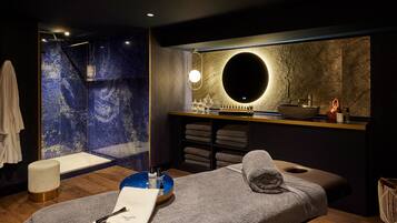 A modern spa room with a marble wall, a round mirror, a wooden bench, and a blue basin.