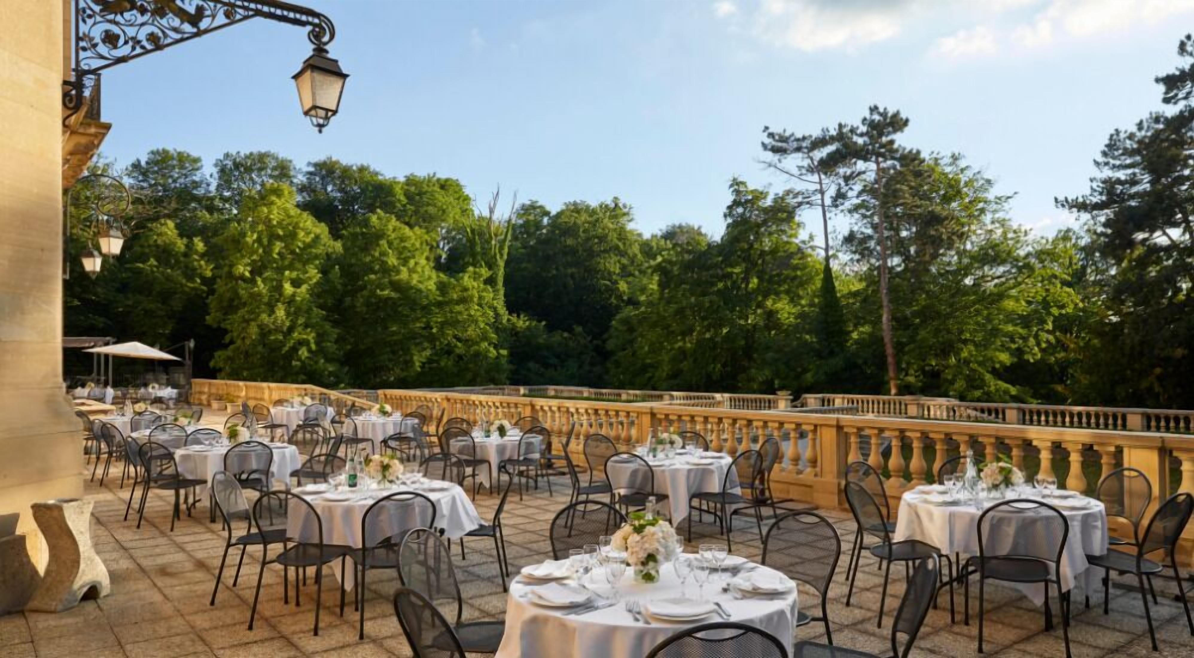 Un área de comedor al aire libre con mesas y sillas redondas, frente a un fondo de árboles y una barandilla de piedra.