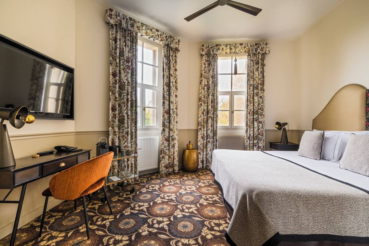 A hotel room with a bed, a desk with a chair, a television, and a large window with curtains.