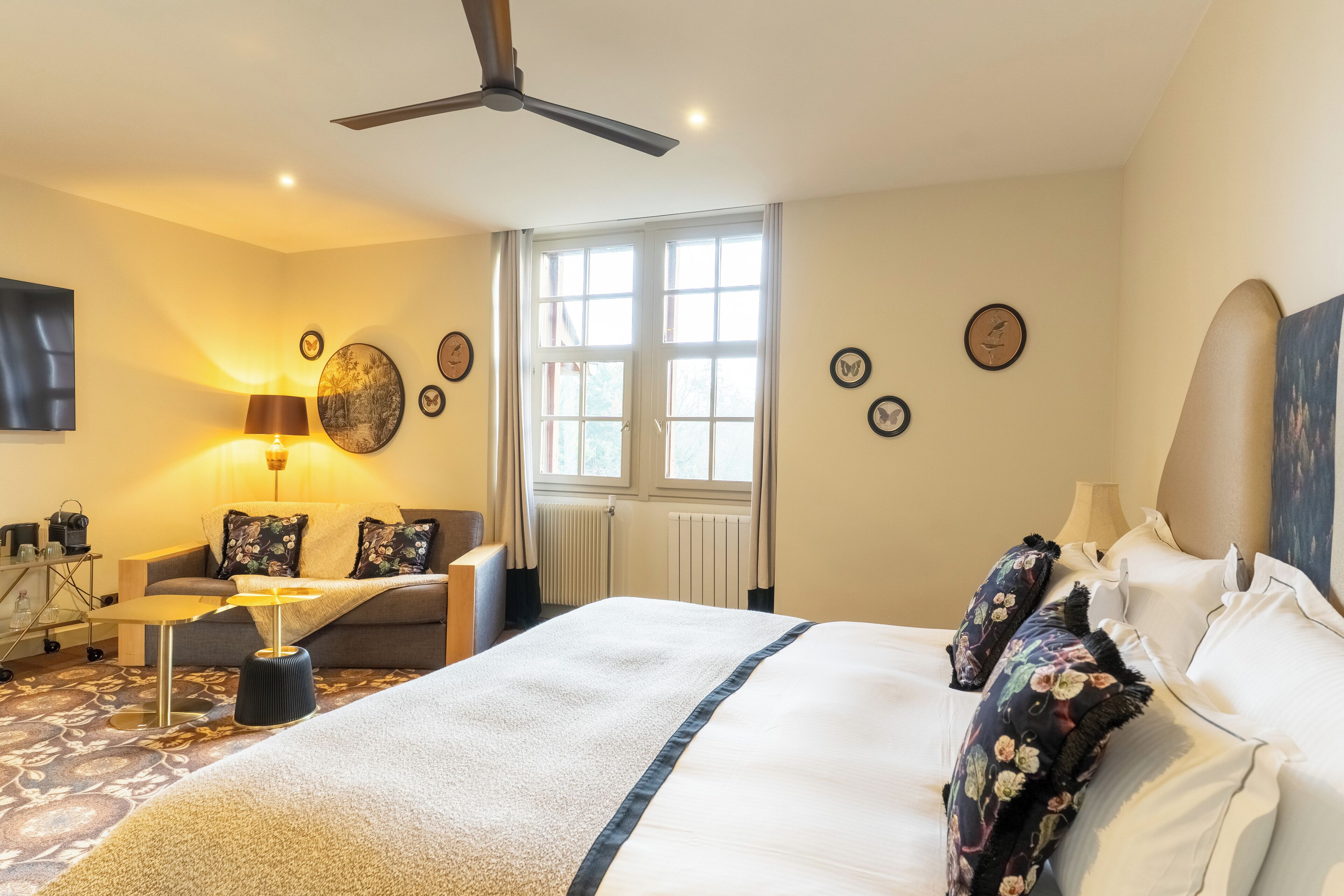 A hotel room with a bed, a desk, a chair, a standing lamp, and a ceiling fan.