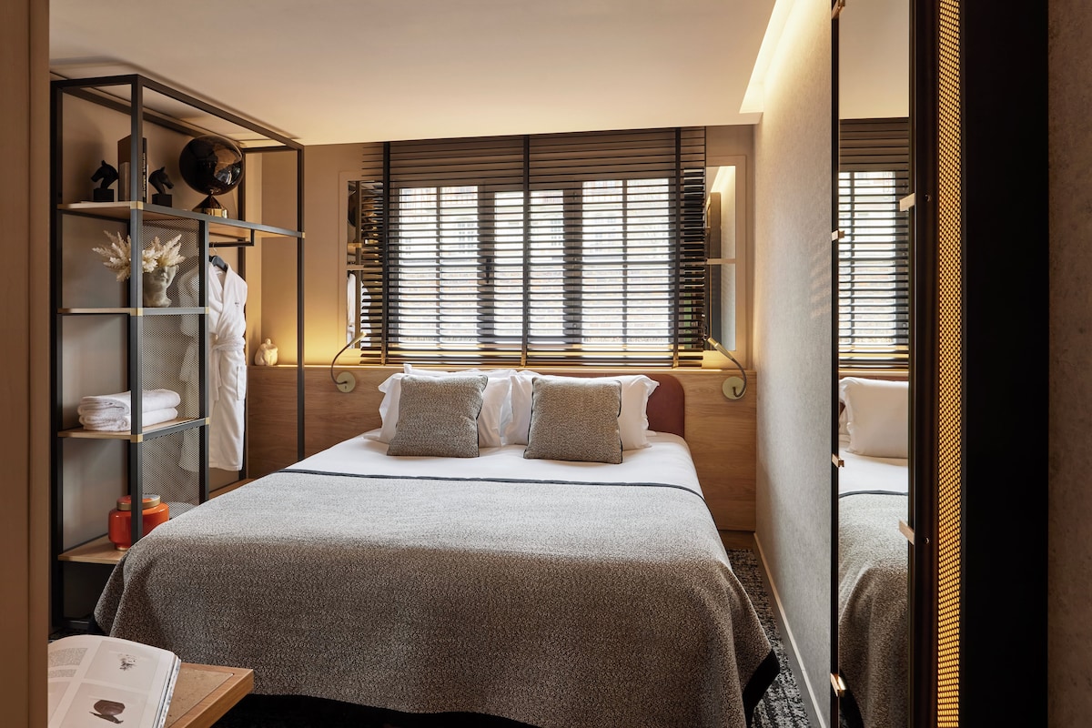 A modern bedroom with a bed, a side table, a bookshelf, and a window with blinds.