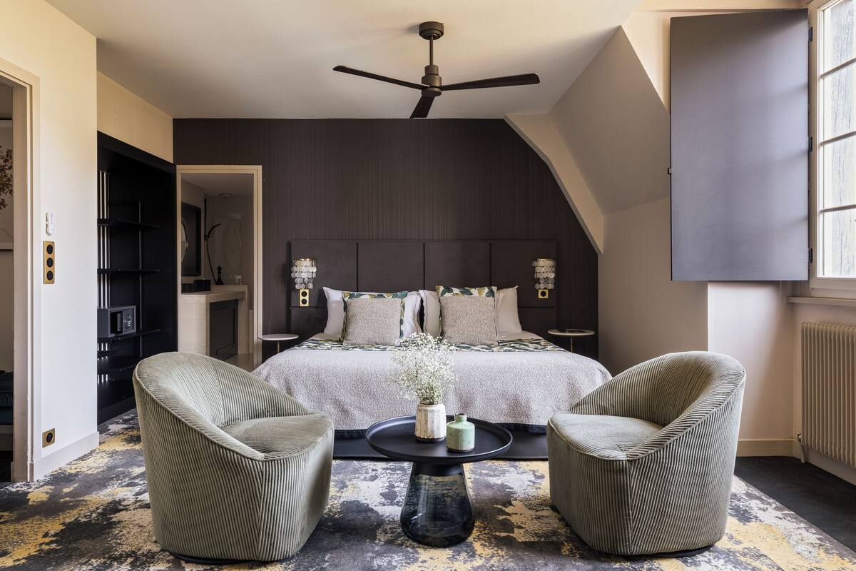 A bedroom with a bed, two armchairs, a coffee table, and a ceiling fan.