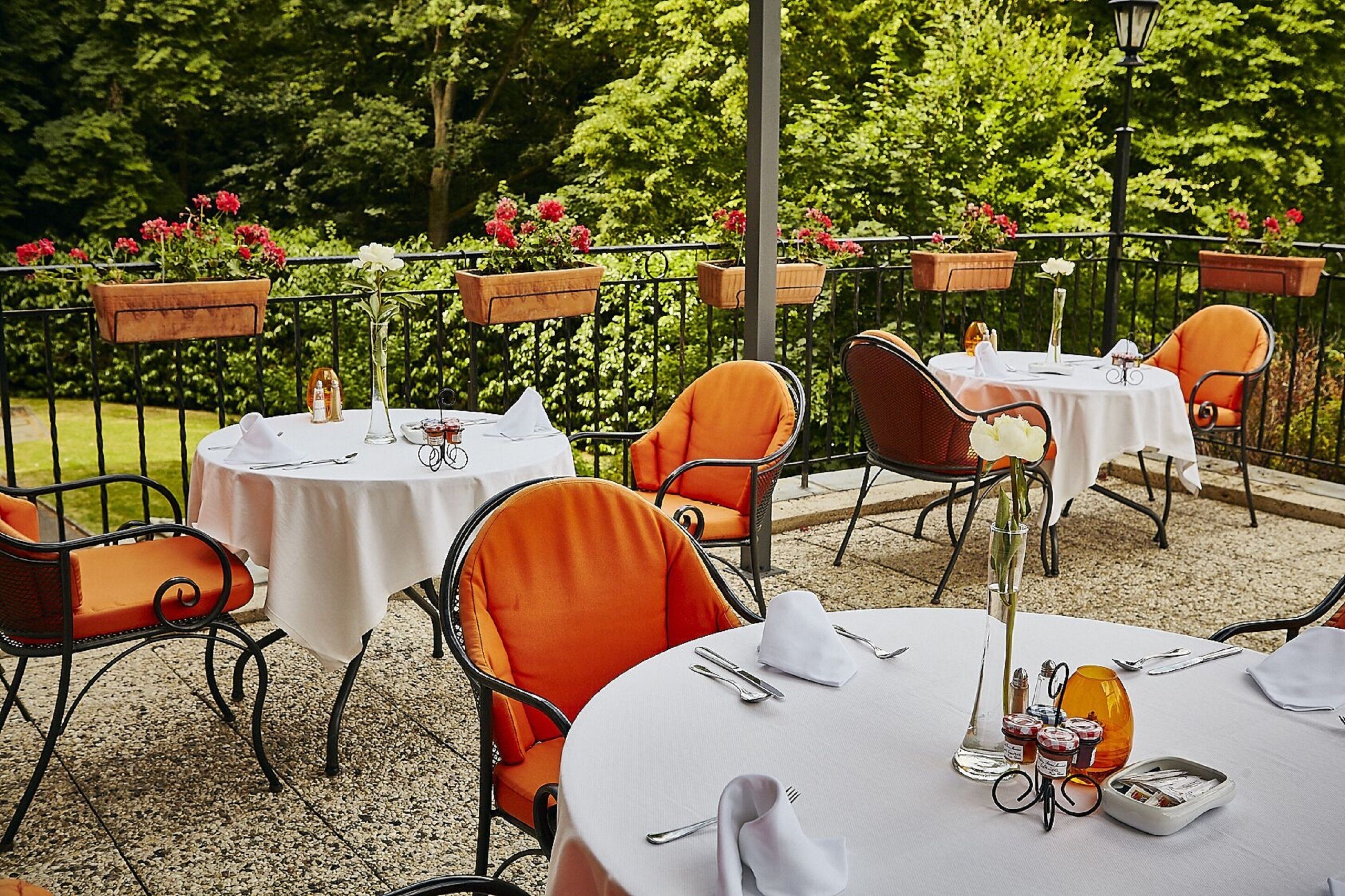 Un espacio de comedor al aire libre con mesas redondas, sillas anaranjadas y flores en macetas.