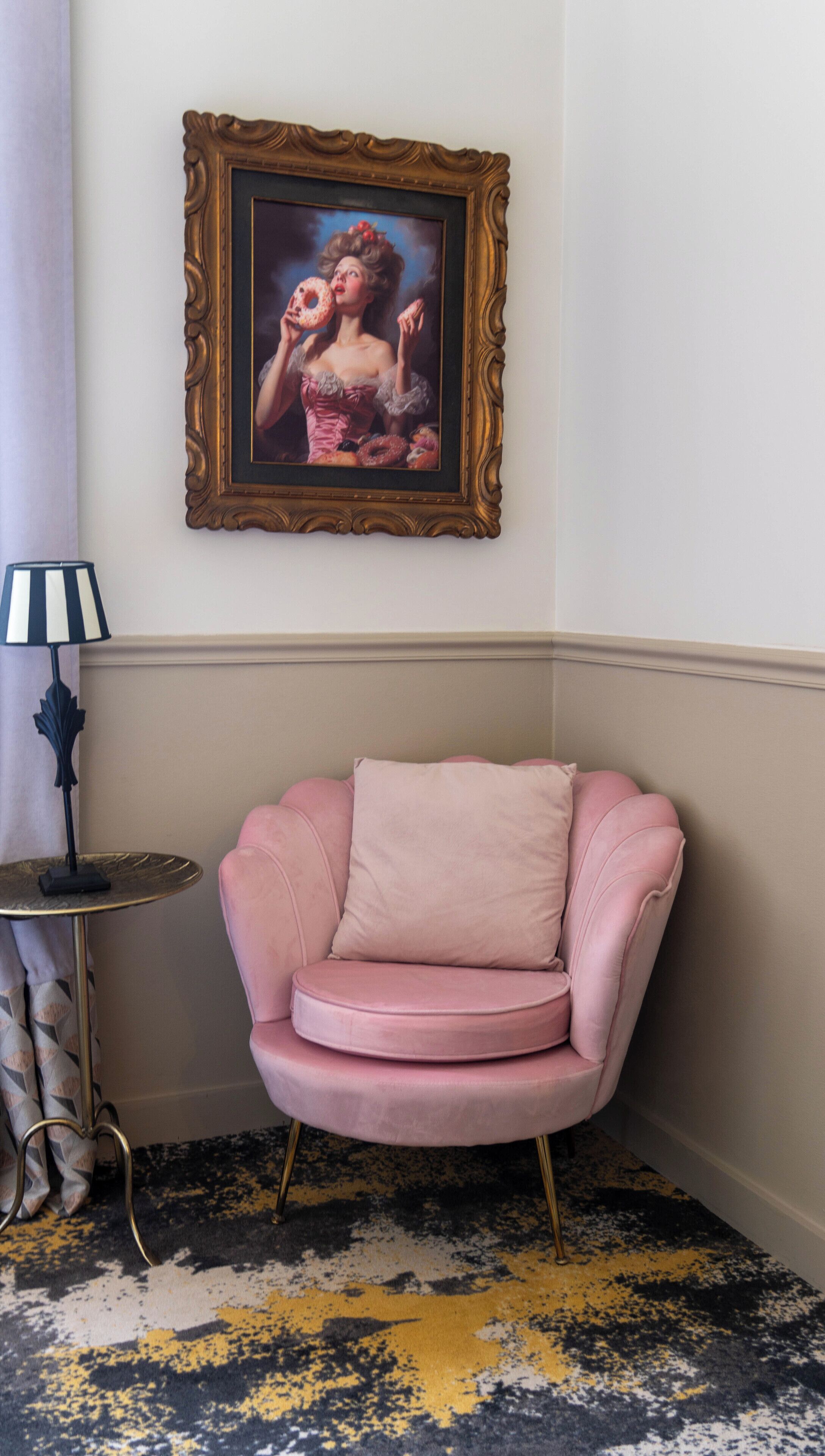 A corner room with a pink armchair, a framed painting, a lamp, and a patterned carpet.