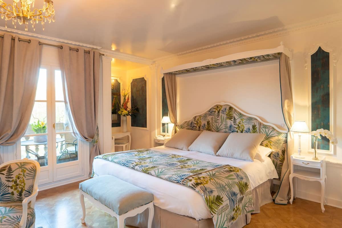 A bedroom with a canopy bed, a sitting area with a chair, a bedside table, and a view of the outdoors.