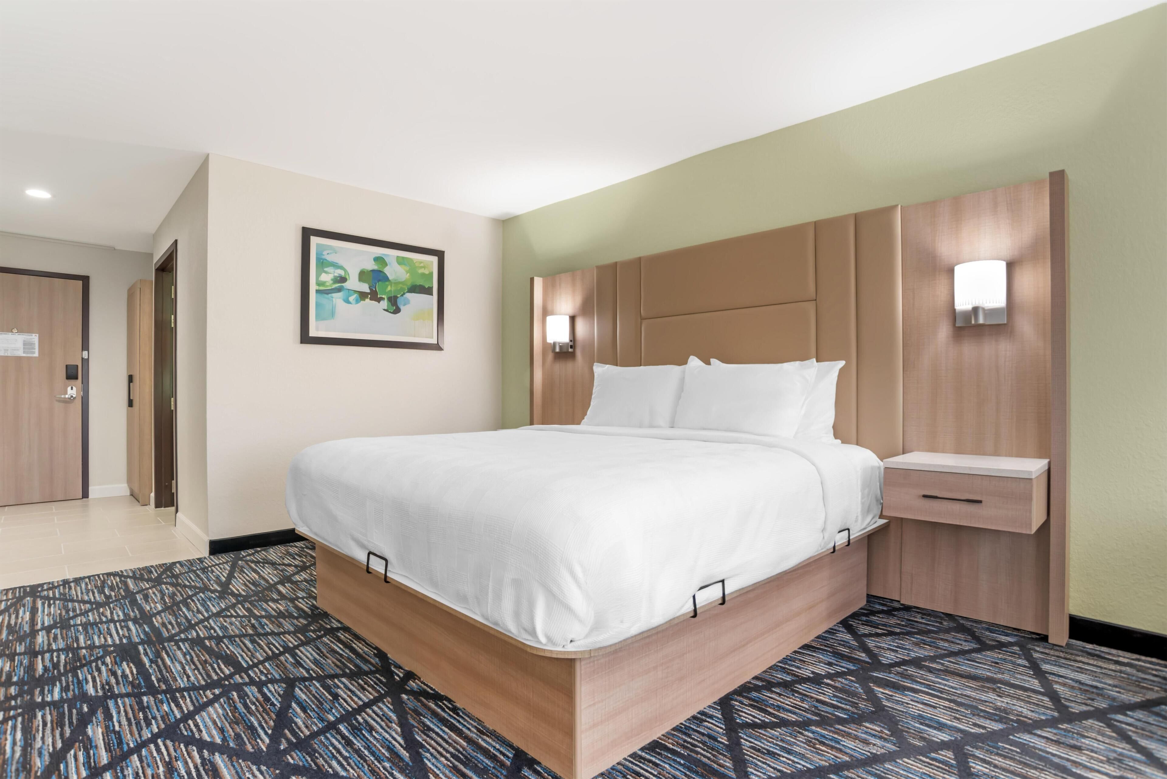Une chambre d’hôtel avec un lit, une table de chevet, une lampe fixée au mur et un tableau encadré accroché au mur.