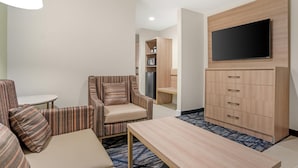 Ein Hotelzimmer mit gestreiftem Ohrensessel, einem kleinen Tisch, einem an einem Holzkasten montierten Fernseher und einem Kühlschrank im Hintergrund.