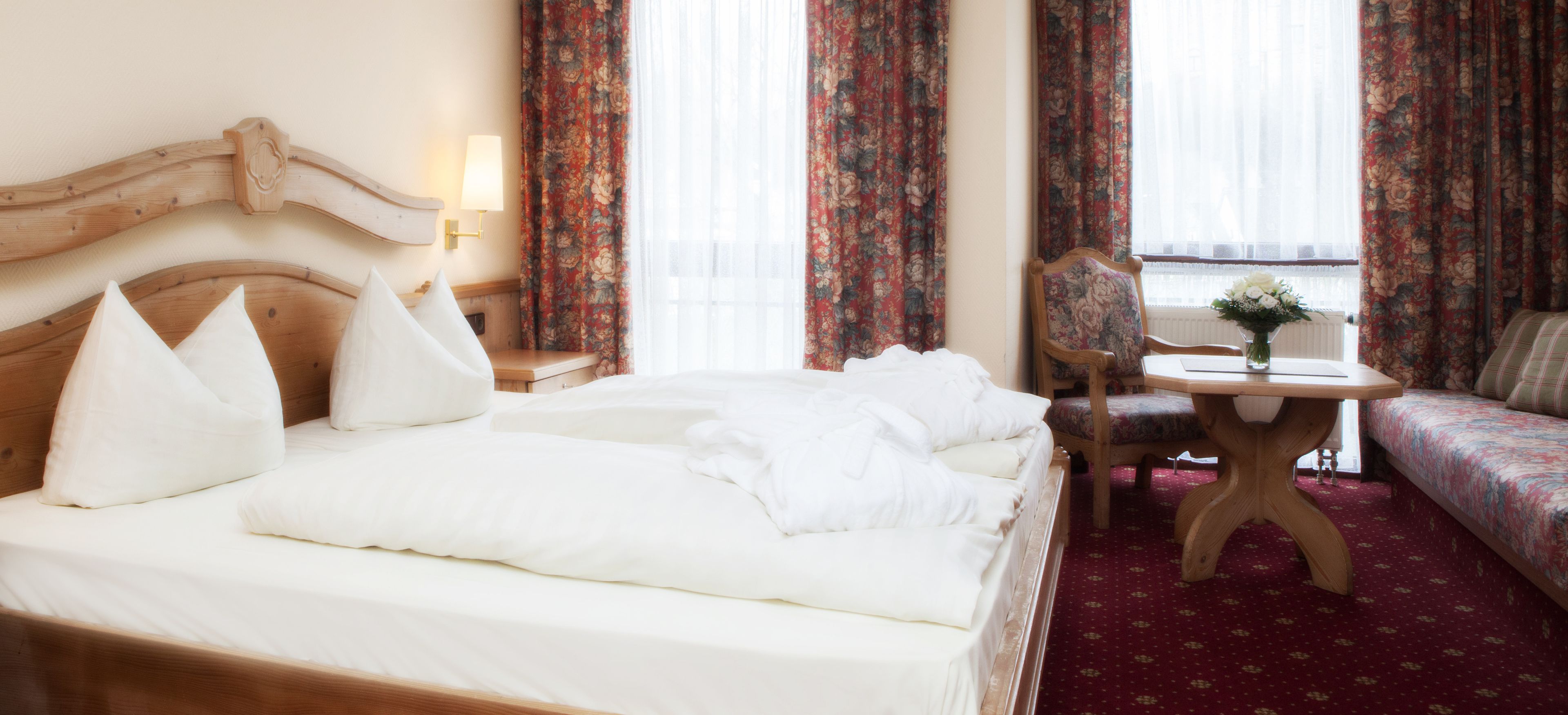 Een hotelkamer met een houten hoofdeinde, een bed met witte lakens, een nachtkastje van hout met een lamp, een bloemenbank en een kleine tafel met een bloevas.