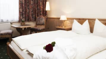A hotel room with a double bed, a wooden headboard, a bedside table with a lamp, a chair, and a small table with a flower vase.