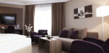 Habitación de hotel con cama, sofá, sillón, una mesita, televisión y un cuadro en la pared.