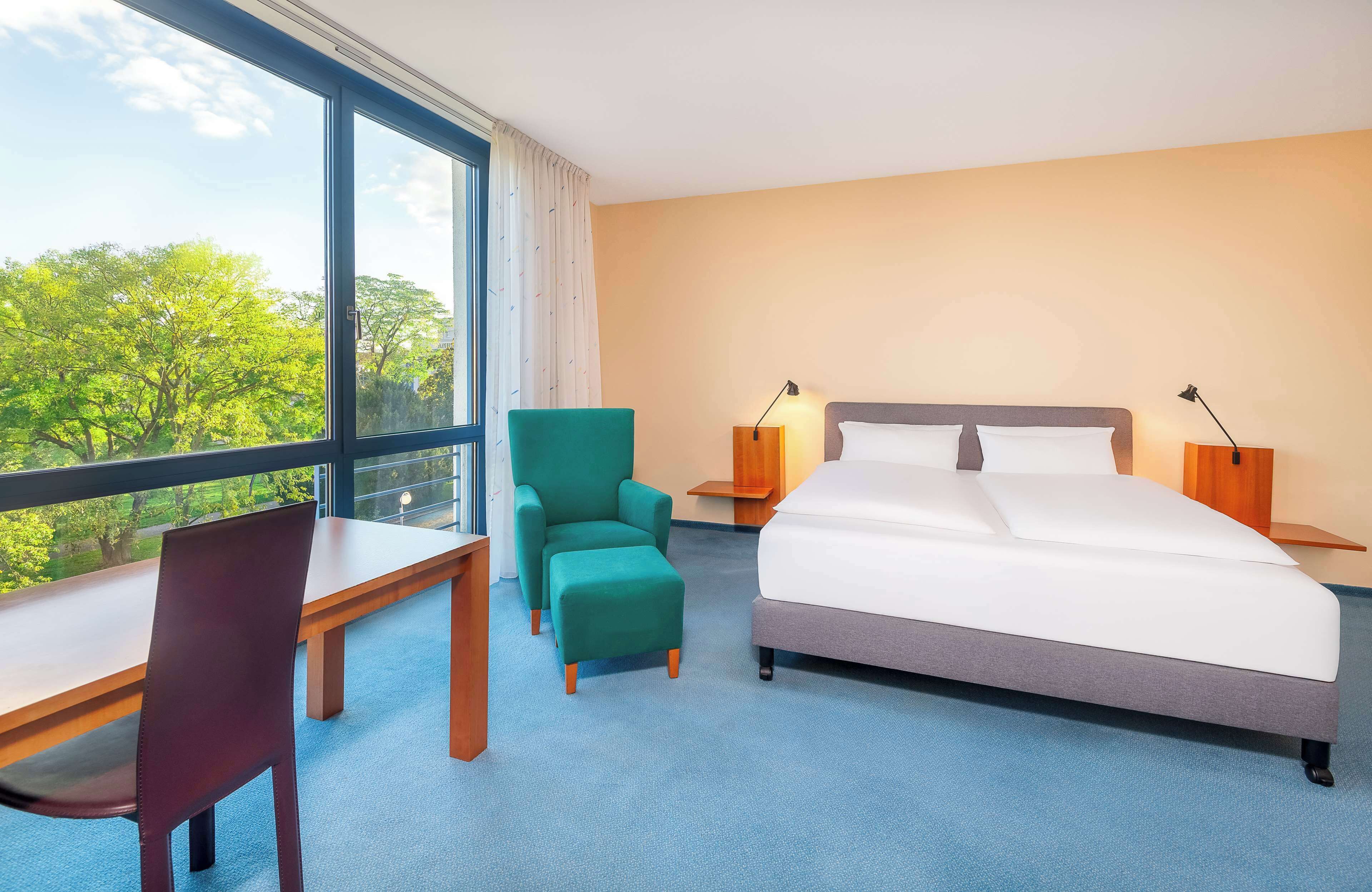 A hotel room with a bed, a desk, a chair, and a view of trees through large windows.
