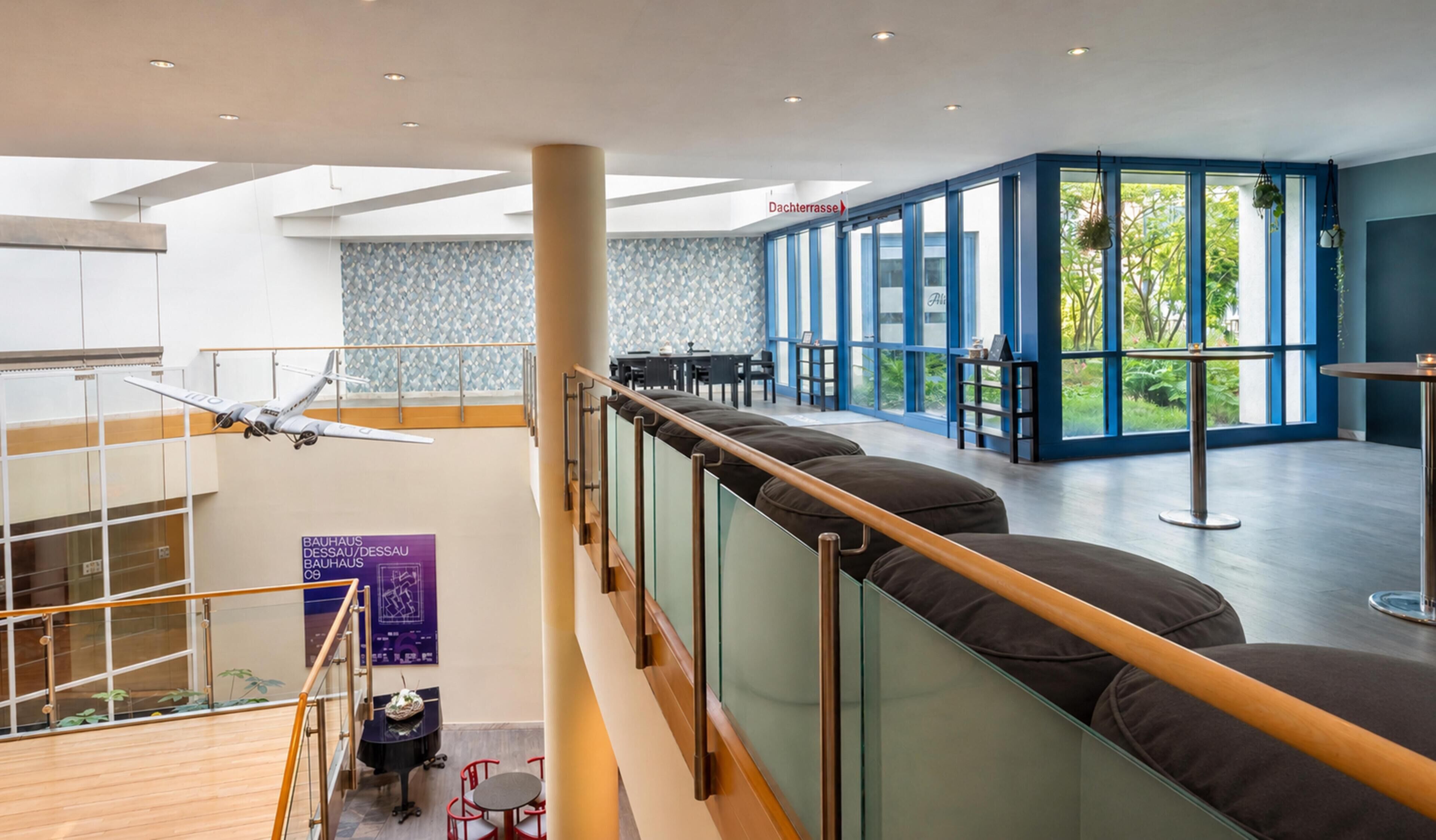 A modern hotel interior with a high ceiling, a staircase, a seating area with cushions, and a view of greenery outside.