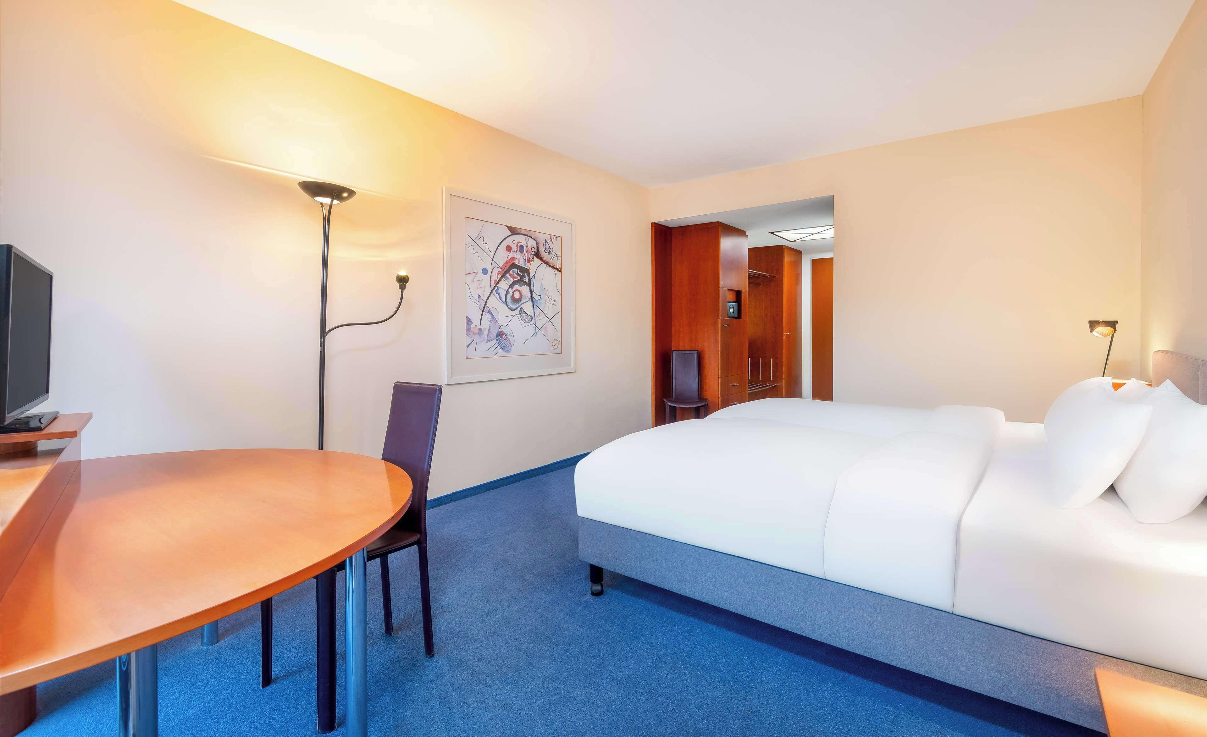 A hotel room with a bed, a desk with a chair, a TV, a painting on the wall, and a door leading to another room.