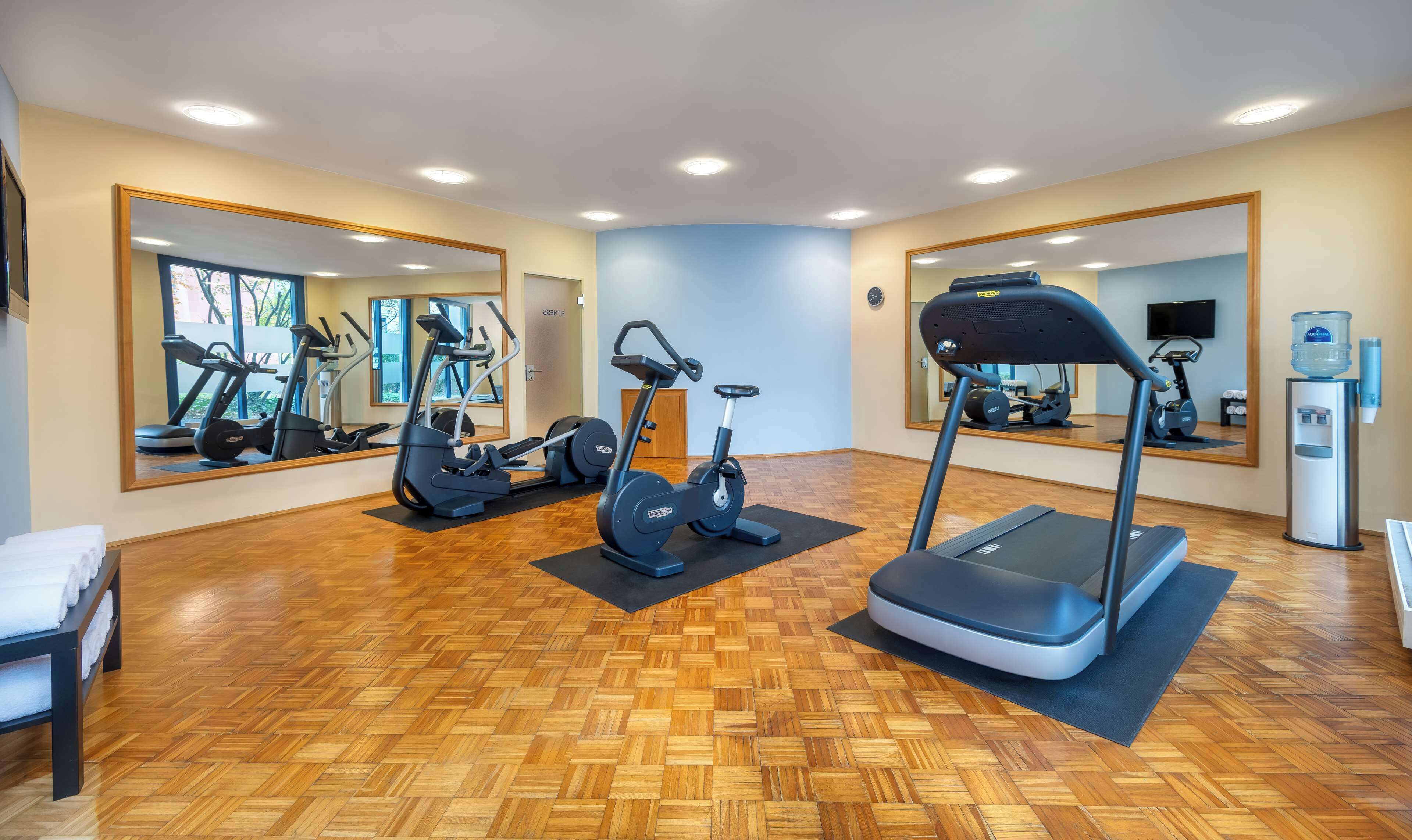 Ein Fitnessraum mit Trainingsgeräten, Spiegeln, Holzboden und einem Wasserspender.
