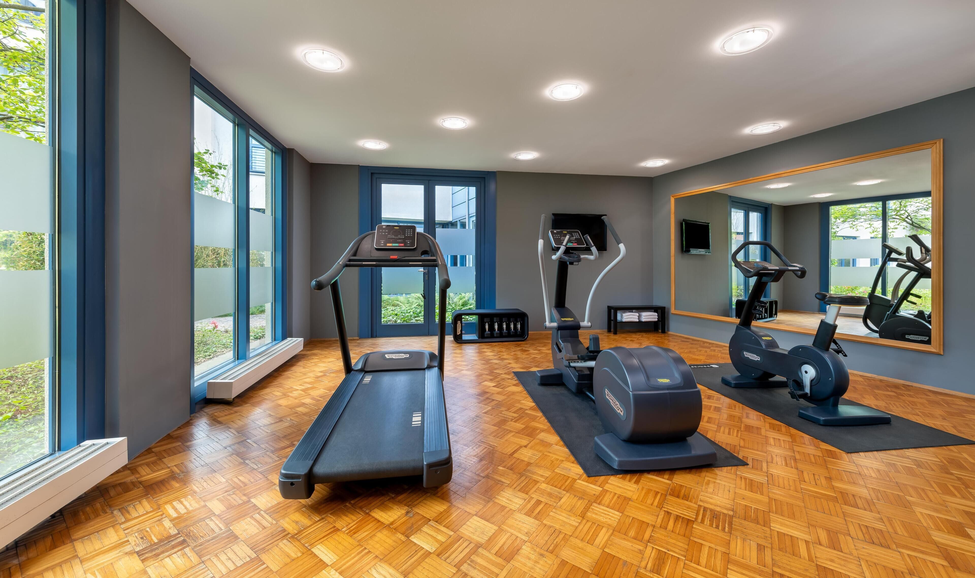 Ein modernes Fitnessstudio mit Laufband, Crosstrainer und Ergometer, großen Fenstern und Spiegeln.