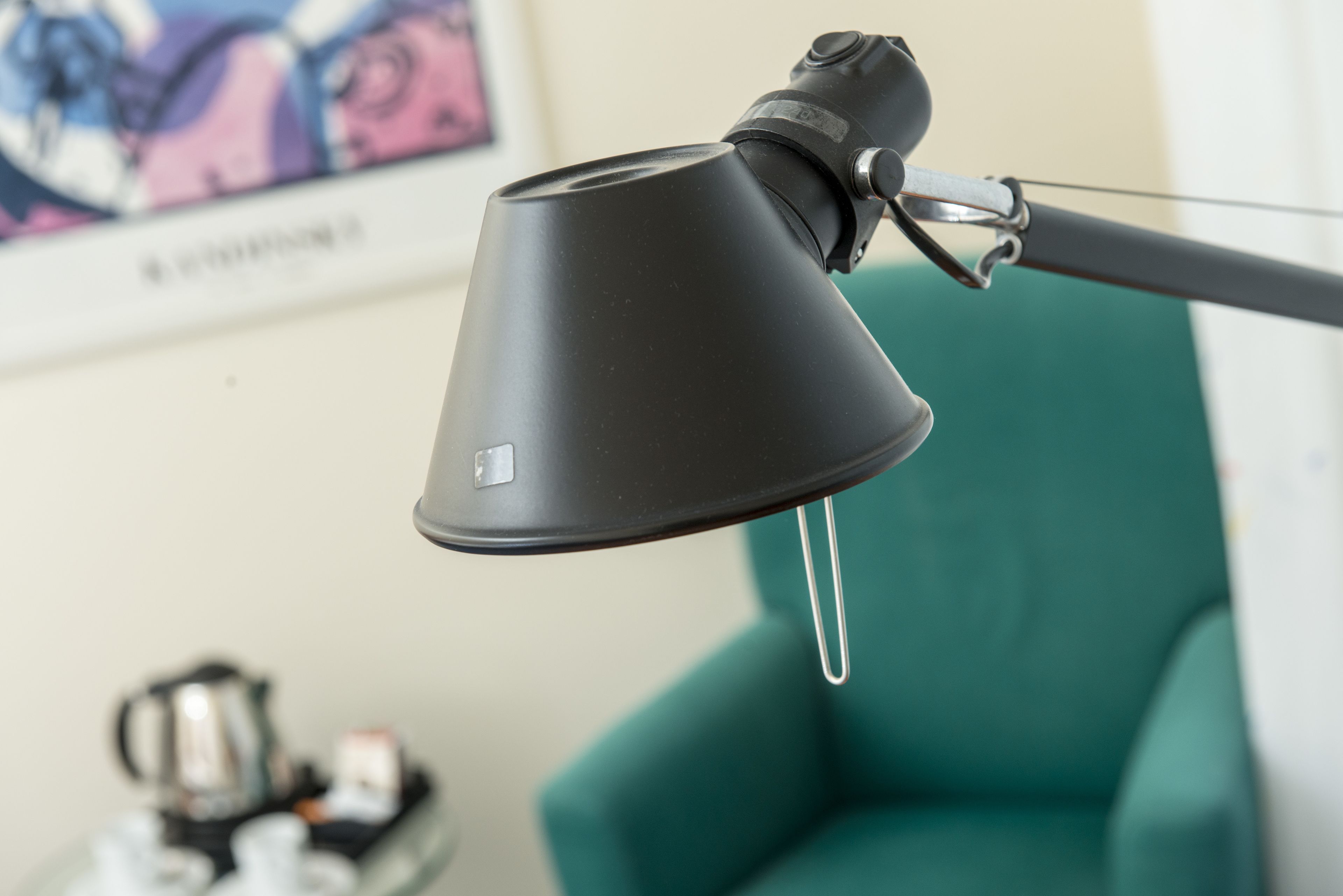 A black desk lamp with a flexible arm, a teal chair, and a small table with a kettle and cups in the background.