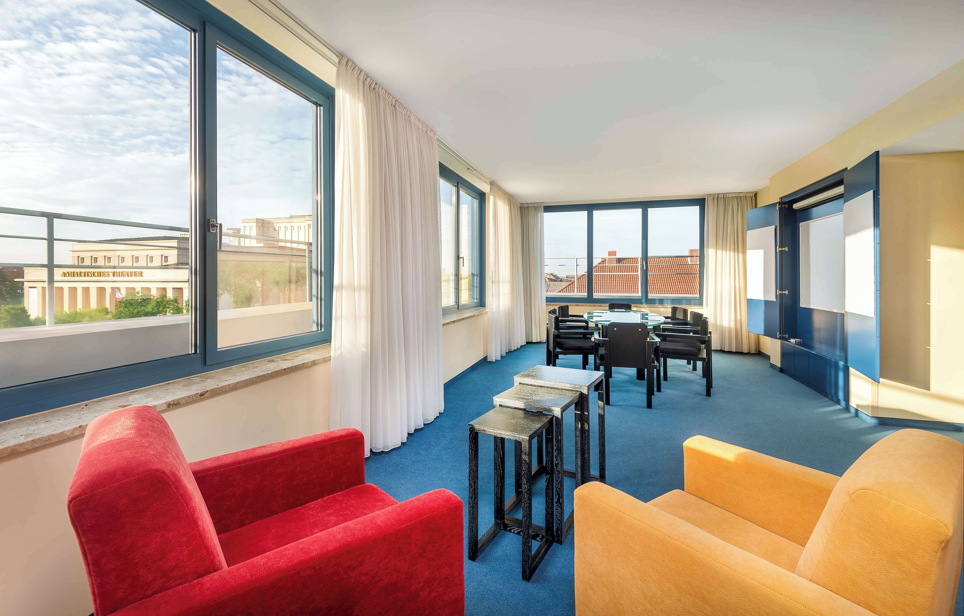 A hotel room with large windows, a balcony, red and yellow chairs, a small table, and a view of buildings.