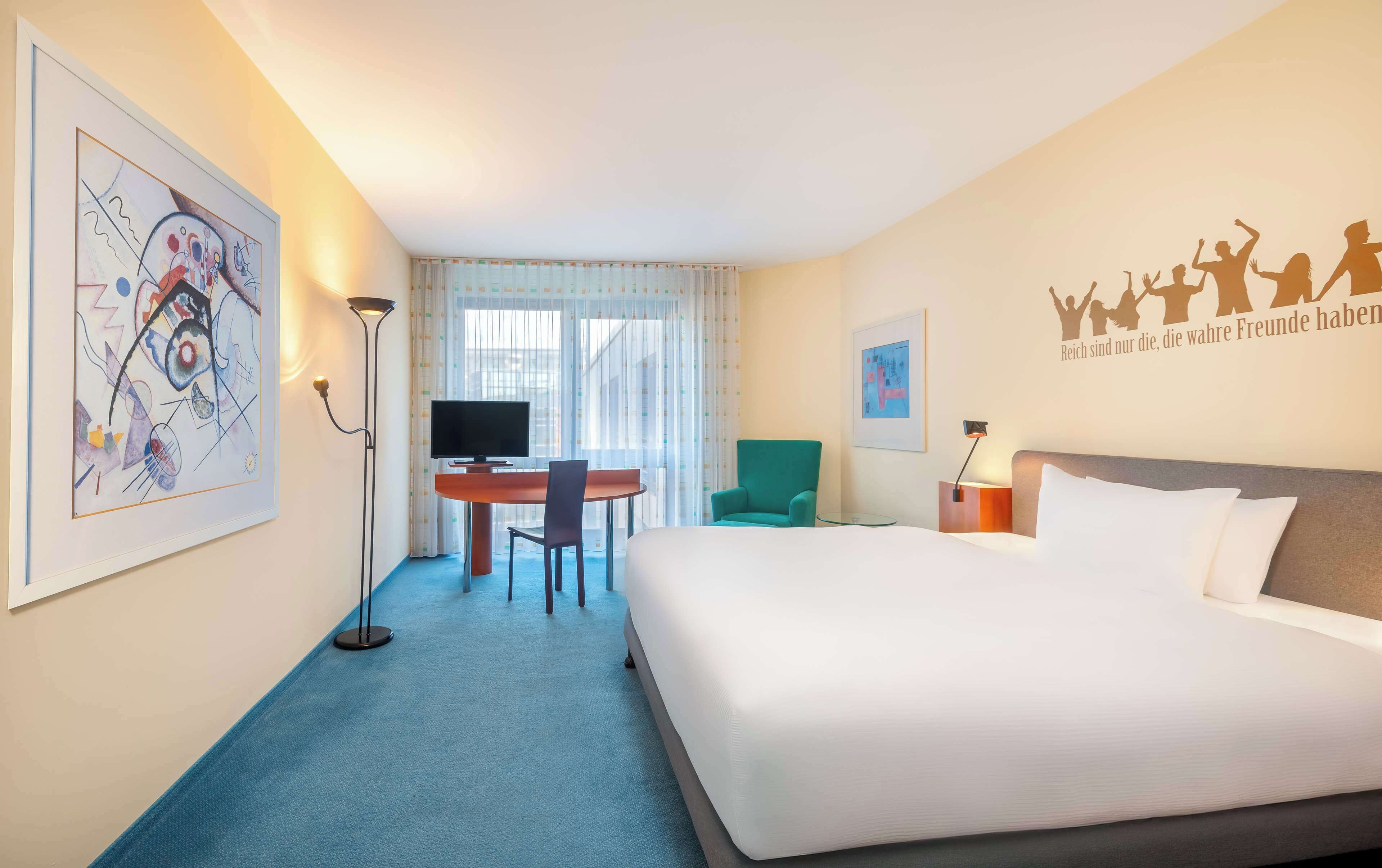 Een hotelkamer met een bed, een bureau met stoel, een televisie en een groot abstract schilderij aan de muur.