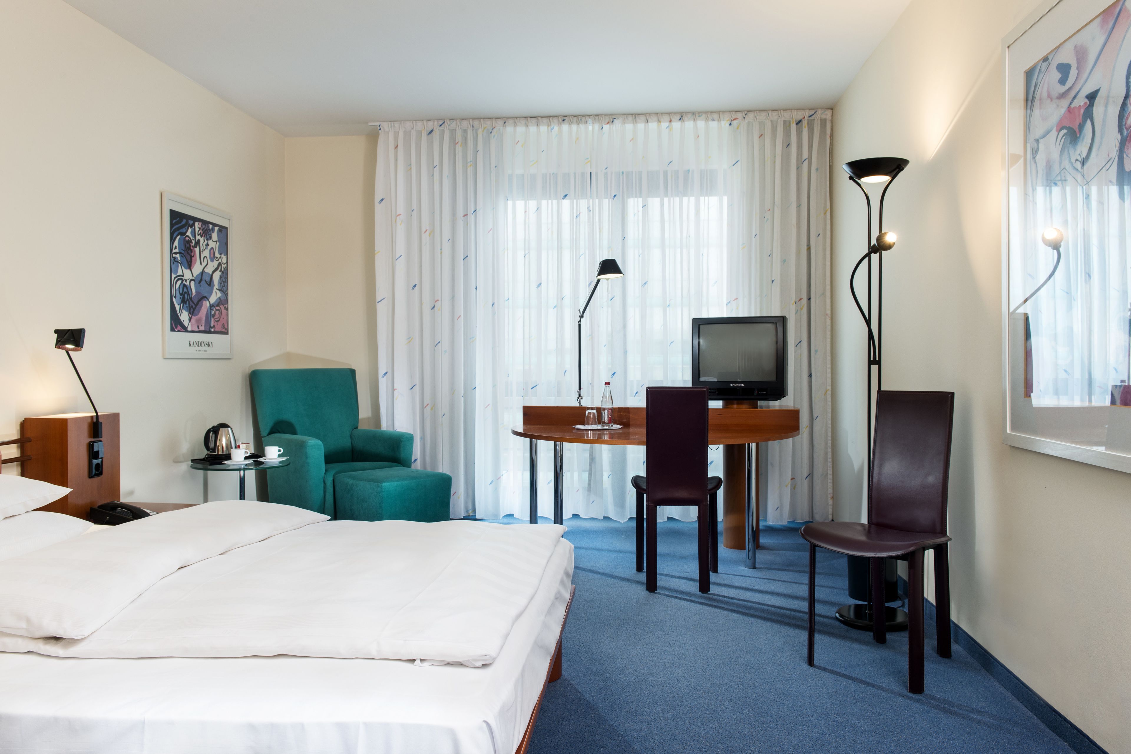 A hotel room with a bed, a desk with a television, two chairs, a lamp, and a painting on the wall.