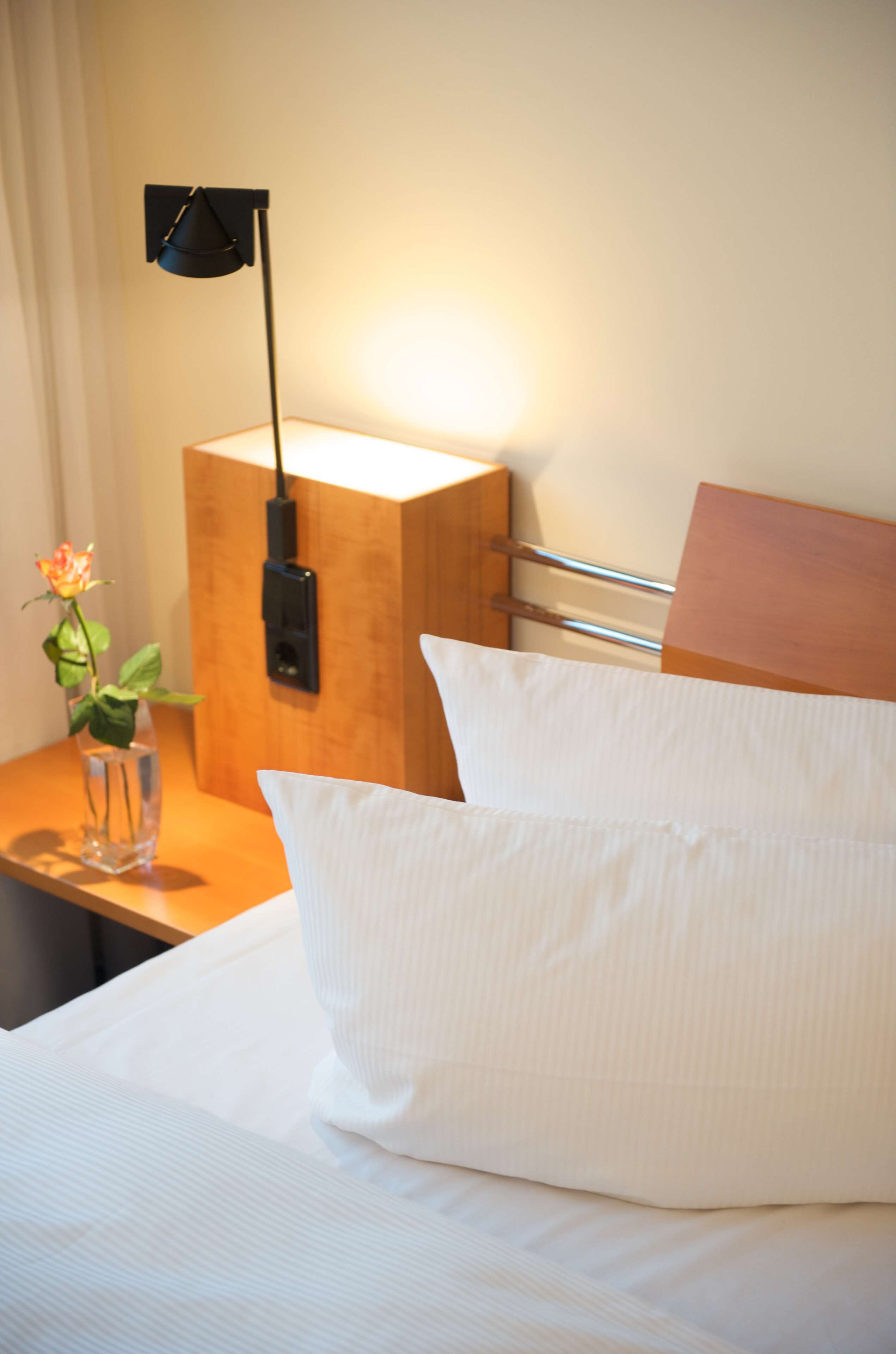 Una cama bien hecha con sábanas blancas, una mesita de noche con un jarrón de flores y una lámpara de pared.