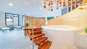 Een modern binnenzwembad met een houten trap naar een jacuzzi, omringd door ligbedden en uitzicht op buiten.