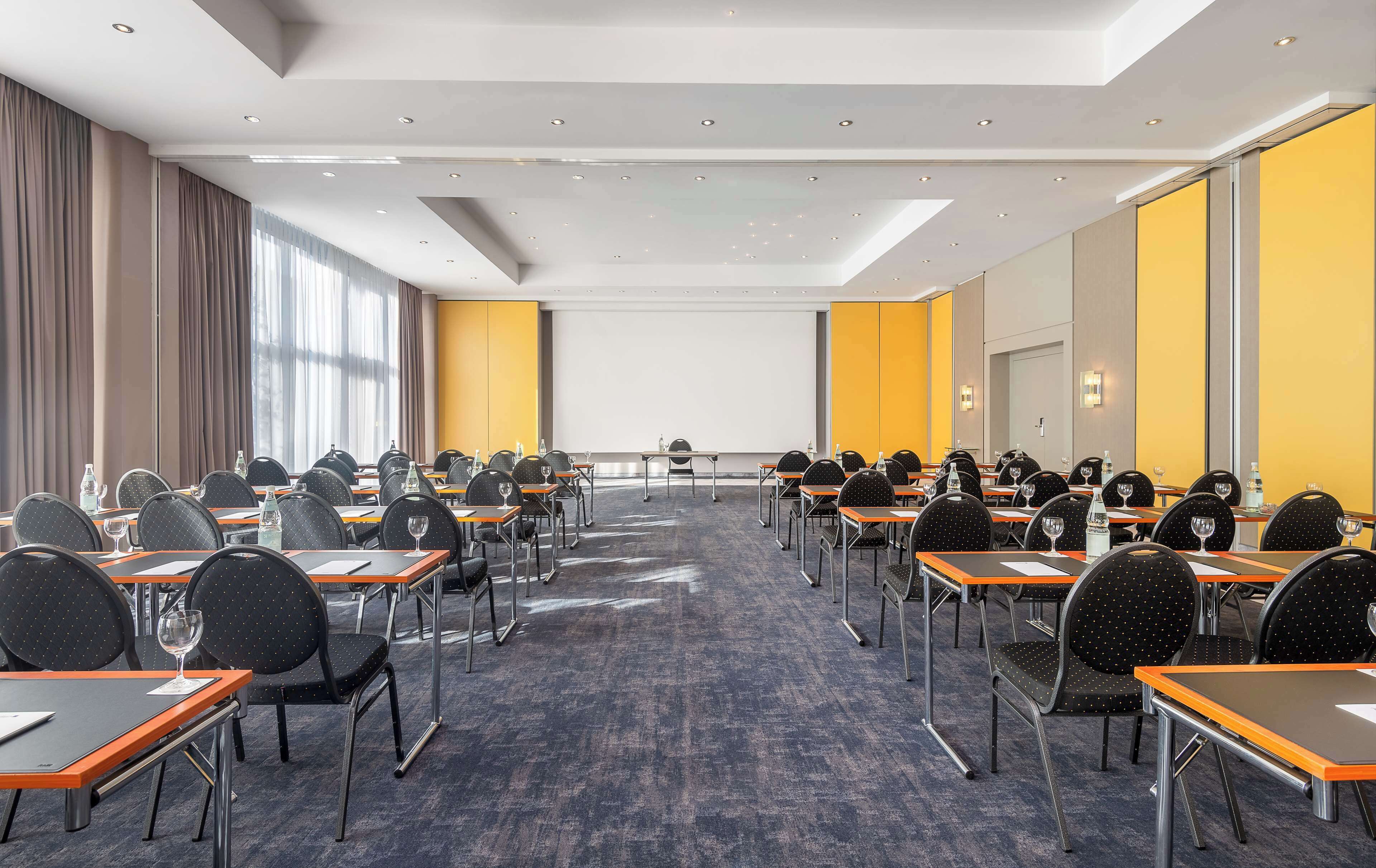 Une salle de conférence avec des rangées de tables et de chaises, de grandes fenêtres et un écran de projection.