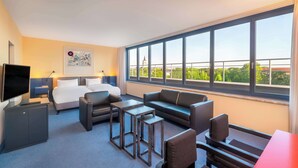 Een hotelkamer met een groot bed, een televisie, een bank, een salontafel en uitzicht op groen en gebouwen door grote ramen.