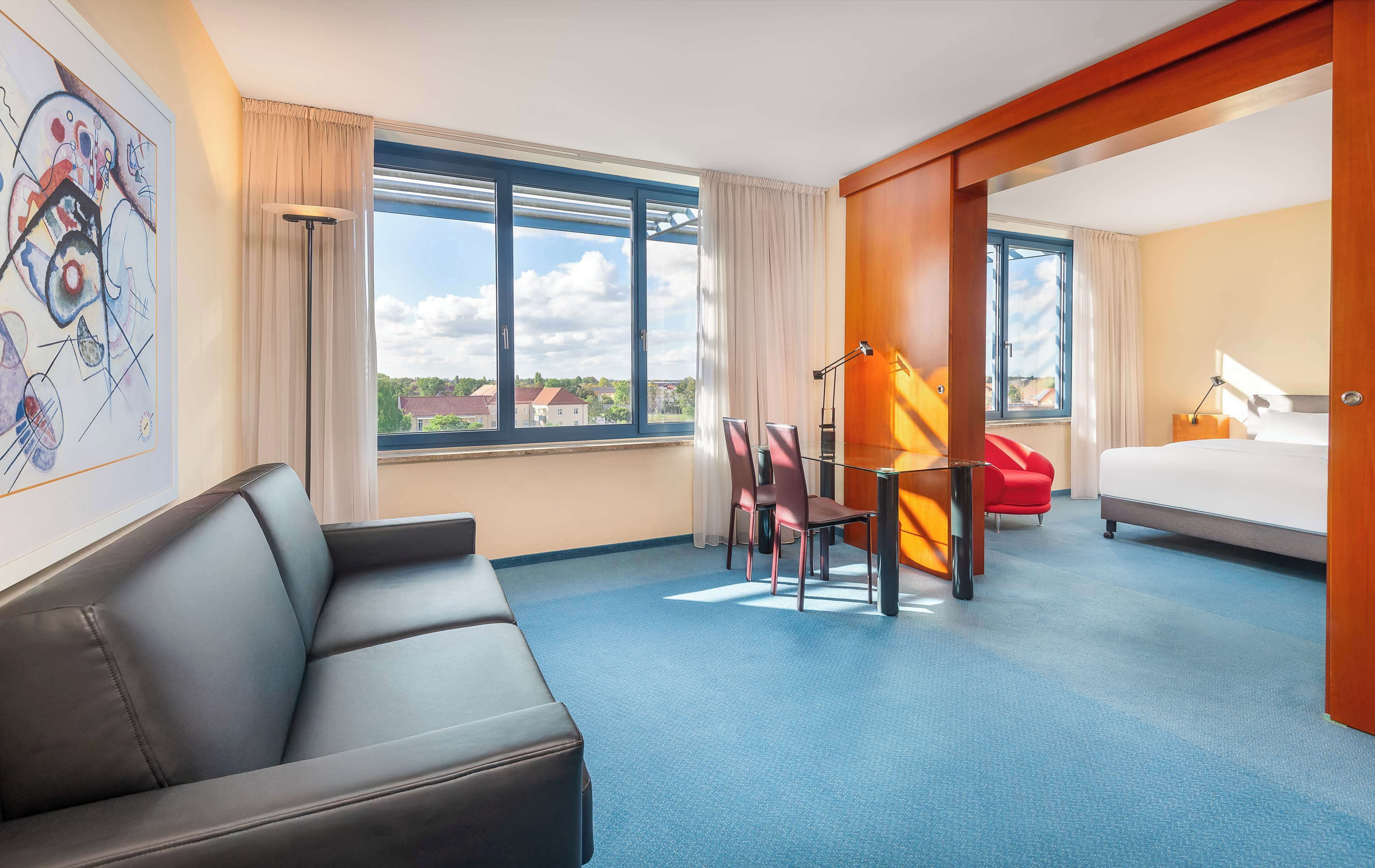 A hotel room with a large window, a bed, a desk with a chair, a sofa, and a painting on the wall.